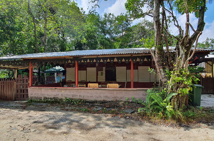 Traditional kampong house at Kampong Lorong Buangkok, featuring a red-pillared open veranda with wooden furniture, set amid dense tropical foliage.