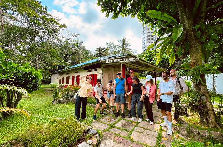 Tour group exploring Kampong Lorong Buangkok, gathered beside a heritage house marked 'Lorong Buong Kua 1954,' surrounded by tropical greenery.