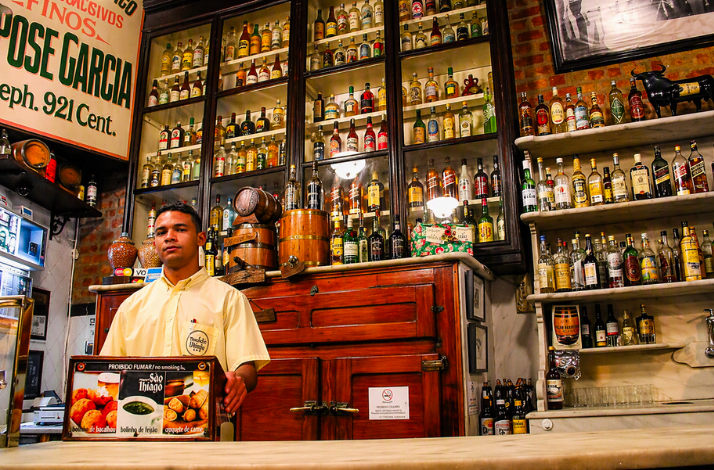 A bartender standing behind a counter in a rustic bar with shelves filled with liquor bottles in Rio.