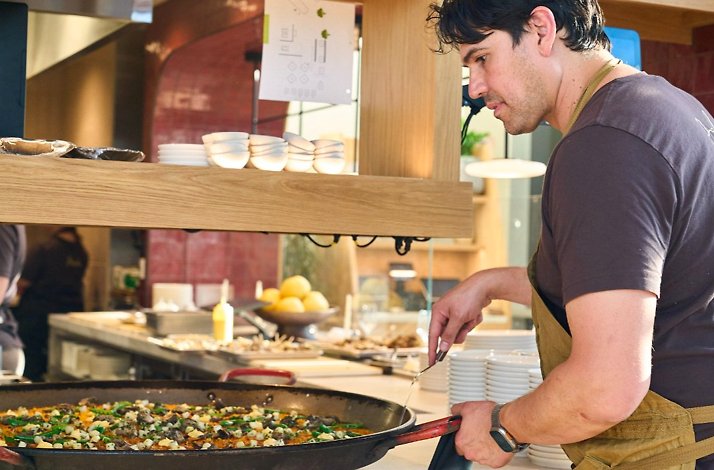 Chef tending a large paella pan at Picala's open kitchen, wearing an apron and focused on the dish.