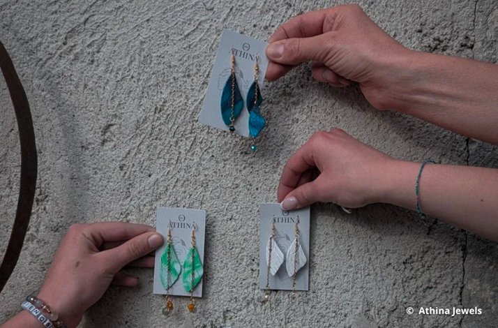 Hands arranging three pairs of handcrafted Athina Jewels leaf-shaped earrings in teal, green, and white displayed on branded cards against a stone wall. © Athina Jewels