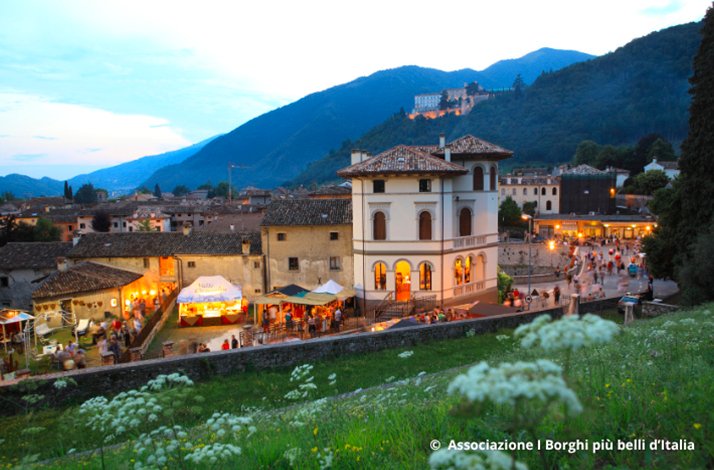 Evening gathering in Cison di Valmarino's historic center, with illuminated market stalls, a white villa, and a floodlit castle on the hillside behind. © Associazione I Borghi più belli d'Italia