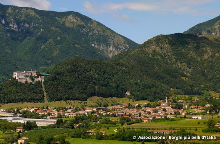Aerial view of Cison di Valmarino, with terracotta-roofed village buildings spreading across a green valley, a hilltop fortress rising on the left. © Associazione I Borghi più belli d'Italia