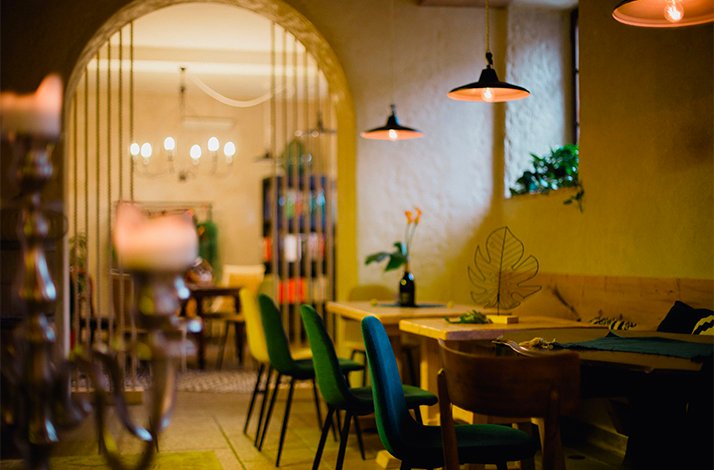 Warmly lit interior of restaurant Vinaria with green velvet chairs, wooden tables, pendant lamps, and a glowing arched doorway opening to an inner room.