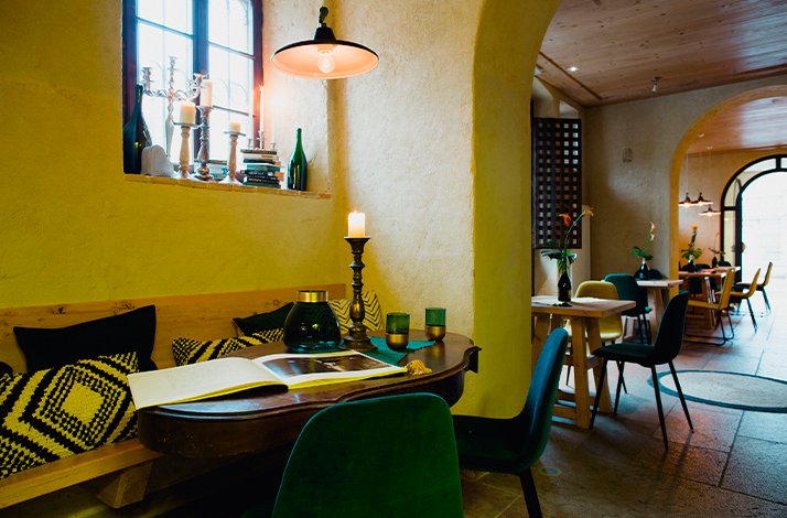 Cozy interior of Vinaria with yellow walls, a candlelit wooden table, open book, green velvet chairs, and arched hallway leading to dining area.