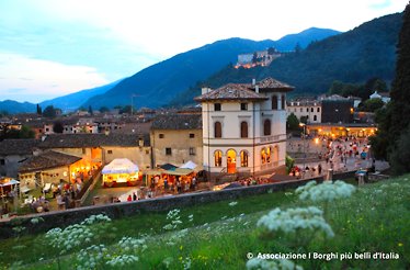 Evening gathering in Cison di Valmarino's historic center, with illuminated market stalls, a white villa, and a floodlit castle on the hillside behind. © Associazione I Borghi più belli d'Italia