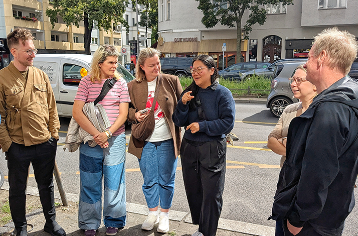 Group of six people chatting on a sunny city sidewalk beside parked cars and apartment buildings.