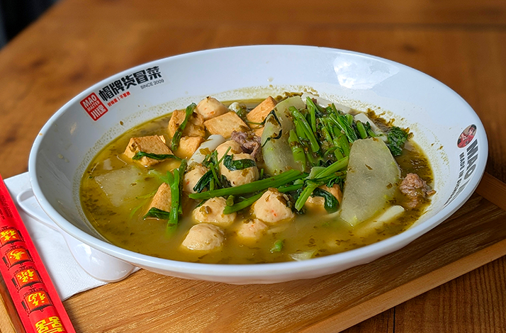 Large bowl of soup with chicken pieces, greens, and radish slices on a wooden table, chopsticks beside it.