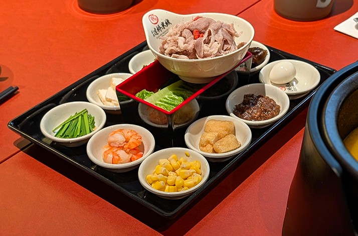 Tray of small bowls with hot pot ingredients—shrimp, corn, tofu, greens, egg, sauces—on a red table.