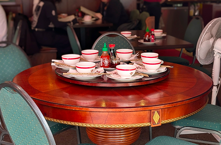 Restaurant table set with bowls and condiments on a lazy Susan, surrounded by green chairs in a dining room.