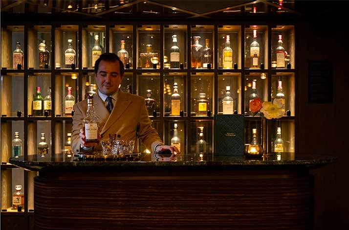 Bartender presenting a whisky bottle at The Londoner's warmly lit bar, surrounded by an extensive backlit whisky collection.