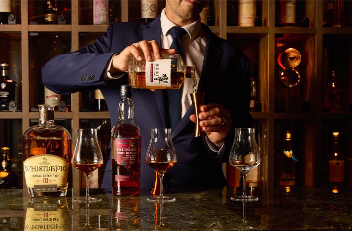 Expert pouring Nikka whisky into a Glencairn glass beside WhistlePig and Talisker Port Ruighe bottles in a warmly lit whisky room.