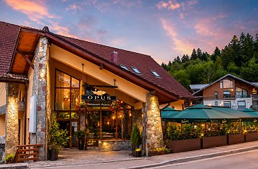 Exterior facade of Opus Garden Restaurant in Sinaia at sunset, with 'Opus Restaurant · Pizzeria' sign, stone columns, lit entrance, and garden terrace seating.