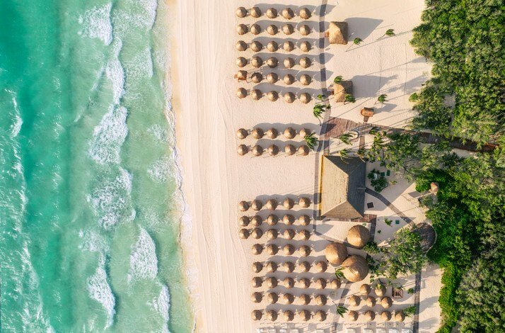 Aerial view of Iberostar Paraiso Lindo's white-sand beach lined with rows of thatched palapa umbrellas beside turquoise Caribbean waves.