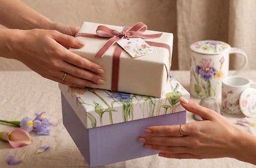 Wrapped gift boxes with satin ribbon being passed between two people, alongside botanical-patterned porcelain mugs, available at Nobila Casa.