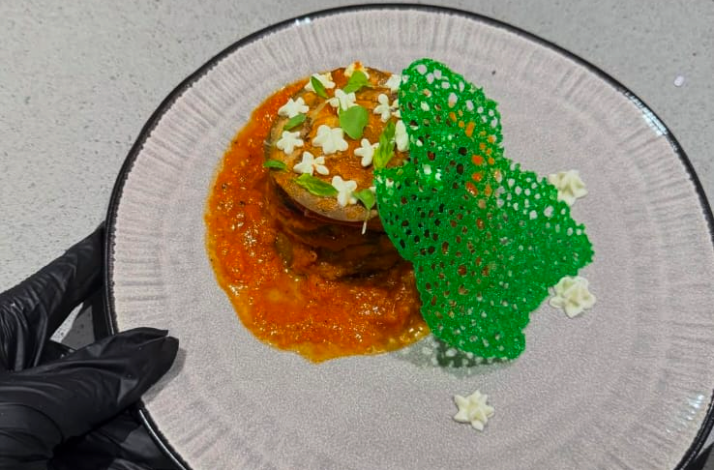 Gloved hands presenting a plated dish of layered ingredients in tomato sauce, topped with herb flowers and a vibrant green lace tuile.