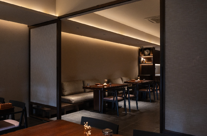 Akar restaurant's intimate dining area viewed through a framed partition, with banquette seating and ambient cove lighting.