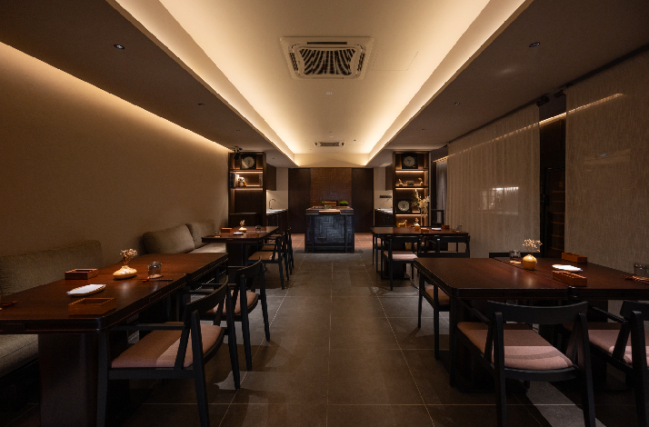 Akar restaurant's softly lit dining room featuring dark wood tables, cushioned chairs, built-in shelving, and warm cove lighting.