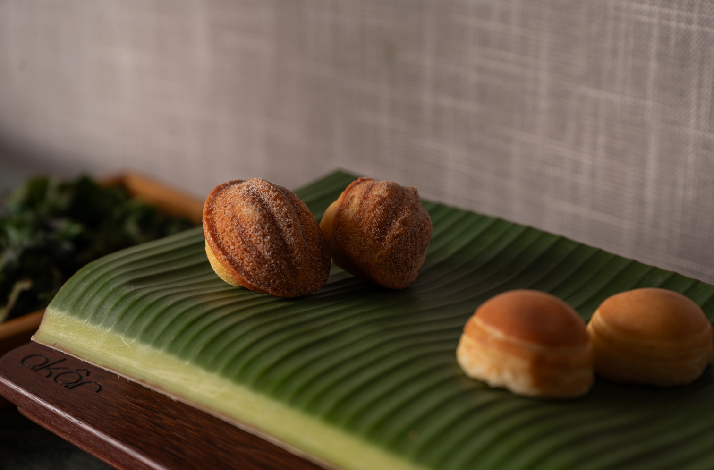 Two spiced buns and two plain rolls rested on a banana leaf atop a wooden board, branded with Akar's logo.
