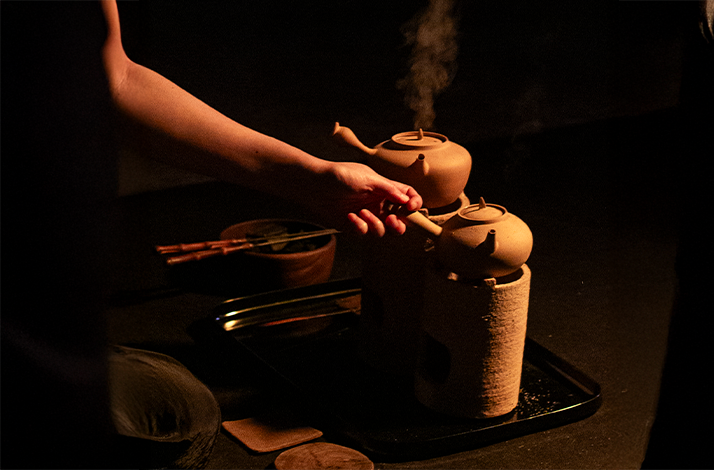 Hands tending stacked clay teapots over a cylindrical heater, with steam rising, during a traditional tea ceremony ritual.