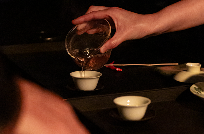 Hands pouring tea from a glass vessel into white ceramic cups arranged on a dark tray during a ceremonial tea service.