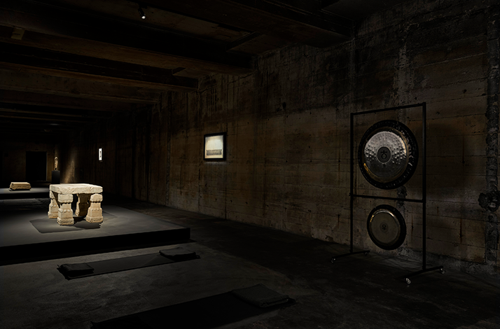 Dim concrete gallery with low platforms, cushions, and a large gong installation on a metal frame at right.