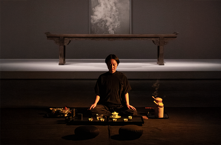 Practitioner seated in meditation before a tea ceremony arrangement, with a Ming-dynasty bench and smoky artwork behind.