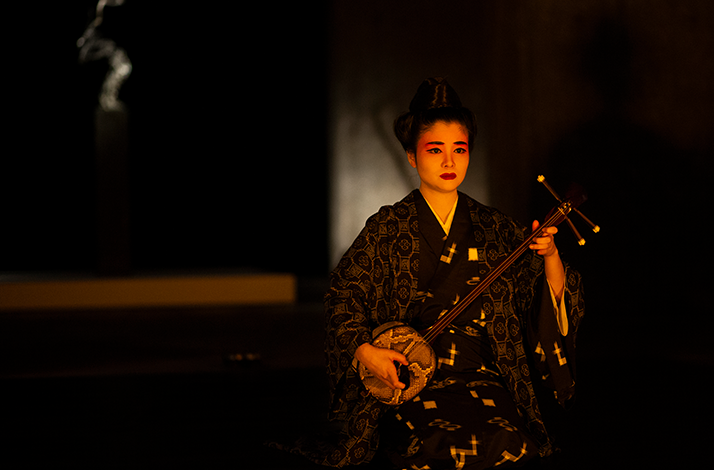 Woman in patterned kimono plays a sanshin under warm spotlight on a dark stage.