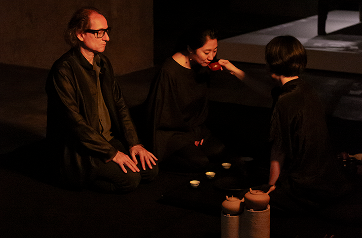 Three guests seated on the floor during a tea ceremony, with a woman sipping from a red teapot served by a tea master.