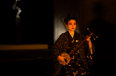 Woman in patterned kimono plays a sanshin under warm spotlight on a dark stage.
