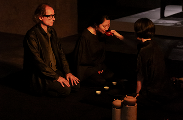 Three guests seated on the floor during a tea ceremony, with a woman sipping from a red teapot served by a tea master.
