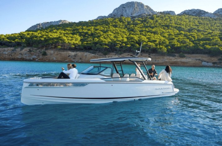 A sleek white motorboat with a few passengers rides on calm turquoise water near a forested shoreline and mountain.