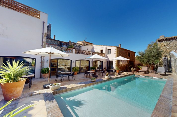 Sunlit courtyard with a turquoise pool, lounge chairs, and white umbrellas outside a rustic stone-and-stucco villa.