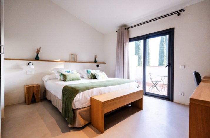 Minimalist bedroom with white bedding and a green throw, wooden bench at the foot of the bed, and sliding glass doors opening to an outdoor terrace.