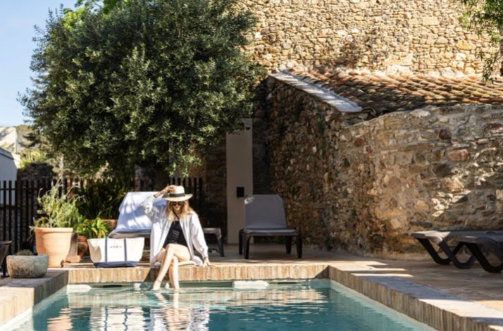 Woman in a sunhat sits on the edge of a pool beside a rustic stone villa.
