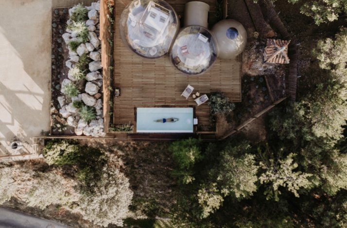 Overhead view of a wooden deck with two transparent bubble suites beside a narrow rectangular pool, surrounded by trees.