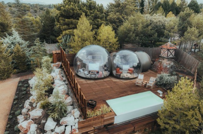 Bubble-style dome rooms on a raised wooden terrace with lounge chairs and a long pool, set in a forested landscape.