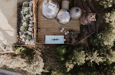 Overhead view of a wooden deck with two transparent bubble suites beside a narrow rectangular pool, surrounded by trees.