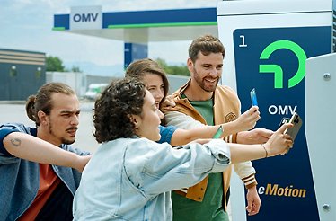 Group of four people interacting with an OMV eMotion EV charger, with an OMV fuel station visible in the background.