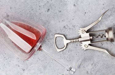 Wine glass filled with rose lying on its side beside a silver winged corkscrew on a gray surface.