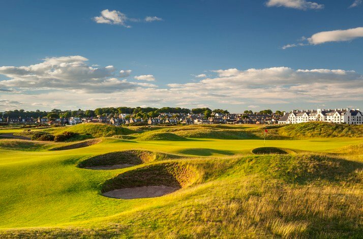Sunlit championship golf hole with undulating green fairways, multiple sand bunkers, and resort buildings on the horizon beneath scattered clouds.
