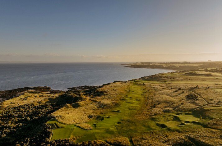 Aerial view of a coastal golf course with a green fairway running along the shoreline at sunset.