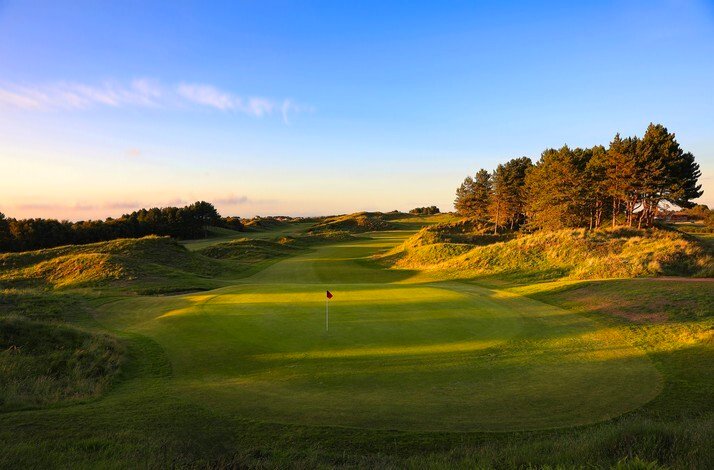 Bunkered links green at Hillside Golf Club bathed in warm evening light, with towering pines and dunes lining the right rough.