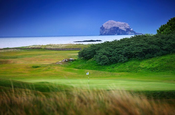 Seaside golf green with a flag on rolling links dunes, overlooking the ocean and a large rocky island under a clear blue sky.