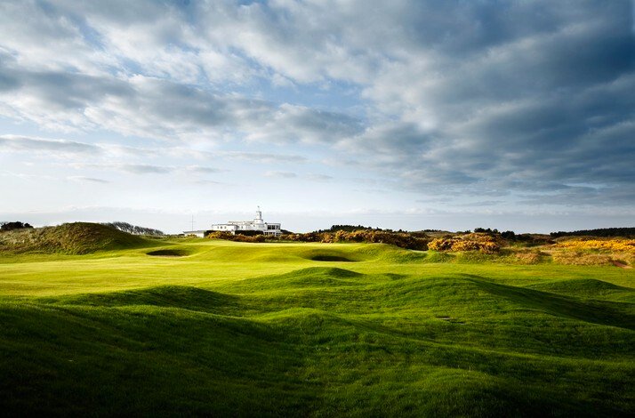 Undulating fairway at Royal Birkdale Golf Club under a moody sky, Art Deco clubhouse and gorse-lined dunes in the background.
