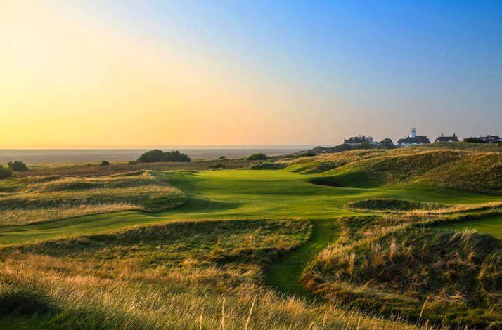 Sweeping links fairway and rough at Royal Liverpool bathed in golden sunset light, coastline and clubhouse in the distance.