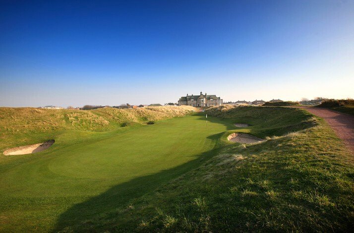Links fairway and bunkered green at Royal Lytham & St Annes under a clear blue sky, clubhouse visible in background. 