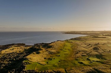 Aerial view of a coastal golf course with a green fairway running along the shoreline at sunset.