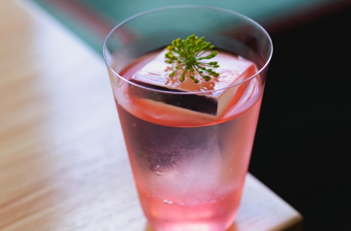Pink cocktail in a clear glass with a large ice cube and a small herb garnish, served at Firstborn.