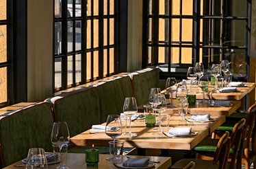 Dining area at Firstborn with sunlit window seating, set wooden tables, and a green banquette.